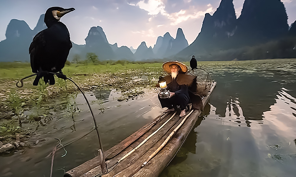 a man sits on a long four log boat in the middle of marshy water surrounded by steep hills and mountains