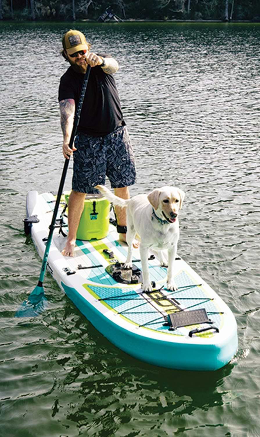 man stands on the Rackham Aero Pedal Drive Paddleboard with his dog, as he paddles with an oar