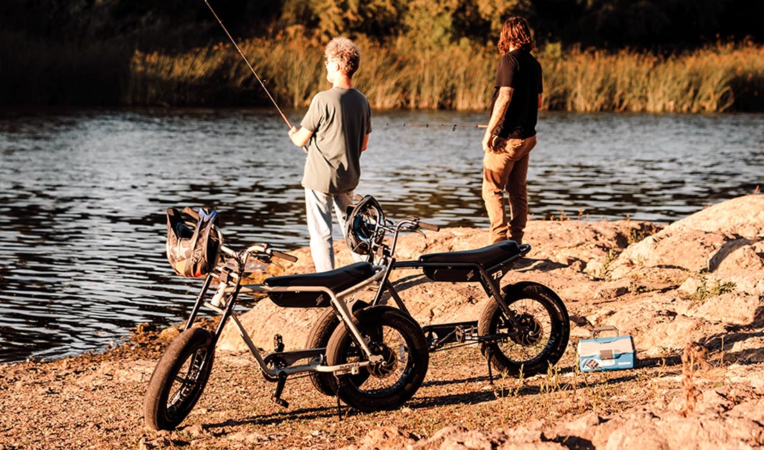 two men fish at a lake with their bikes standing behind them