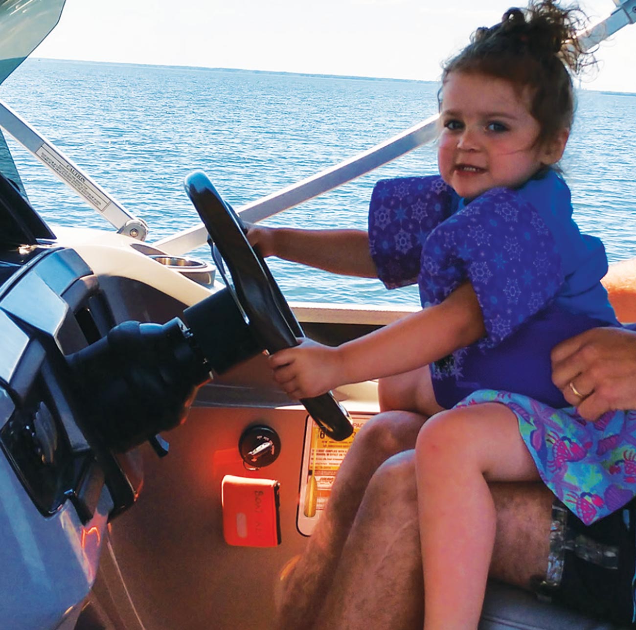 a little girl sits on a man's lap at the wheel of a pontoon