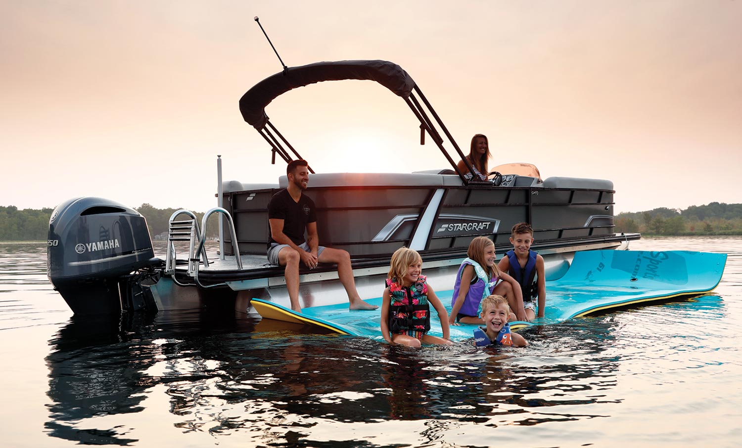 four children on an Aqua Lily Pad floating foam island while two adults watch from a pontoon beside the pad