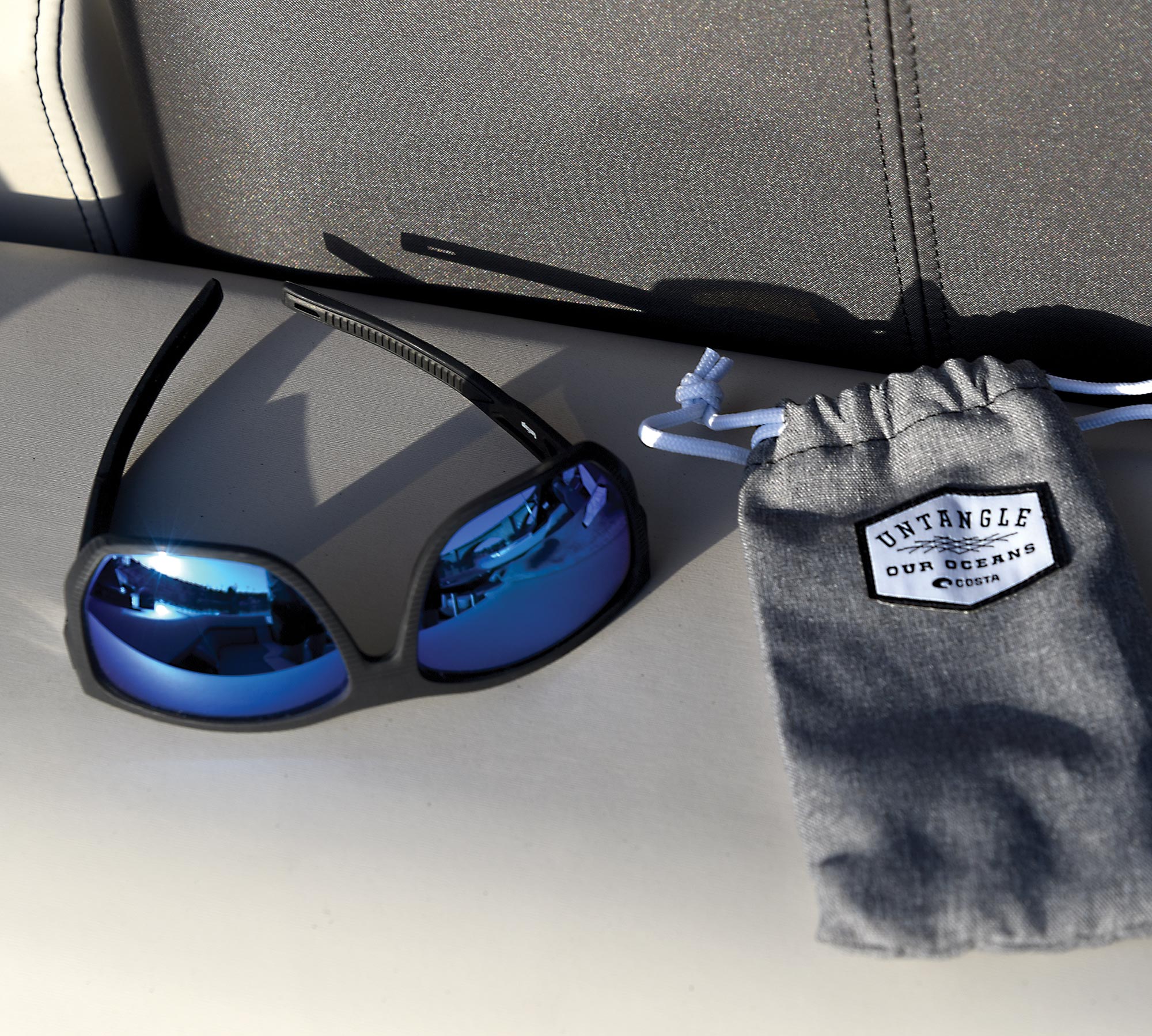 Costa Del Mar Santiago Untangled Sunglasses (blue tinted inner frame and black outer frame) sit upside down next to its grey protective cover case