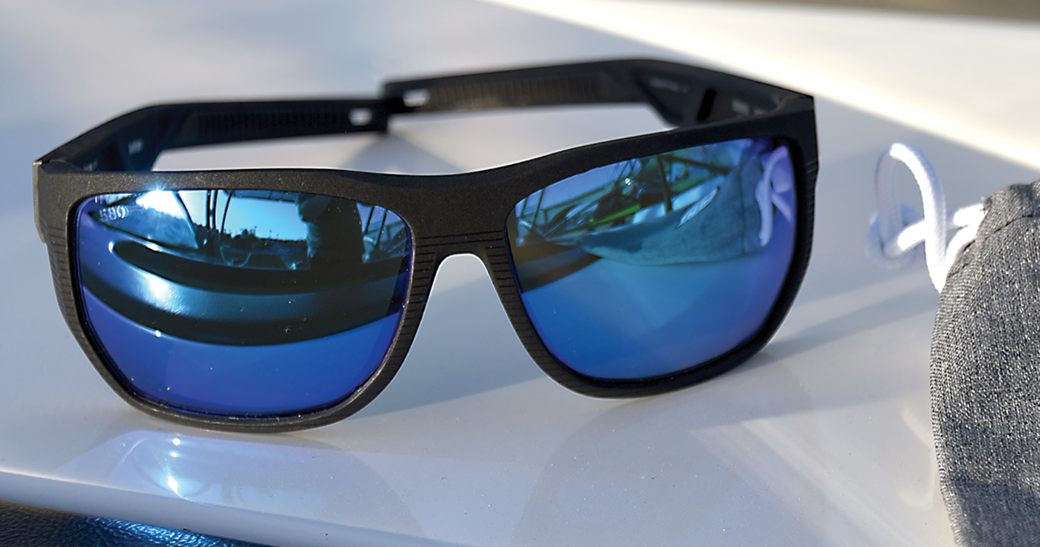 Close-up perspective of the Costa Del Mar Santiago Untangled Sunglasses (blue tinted inner frame and black outer frame) with one part of the inner frame shining a blue light on the surface from the sun's beam/shadow