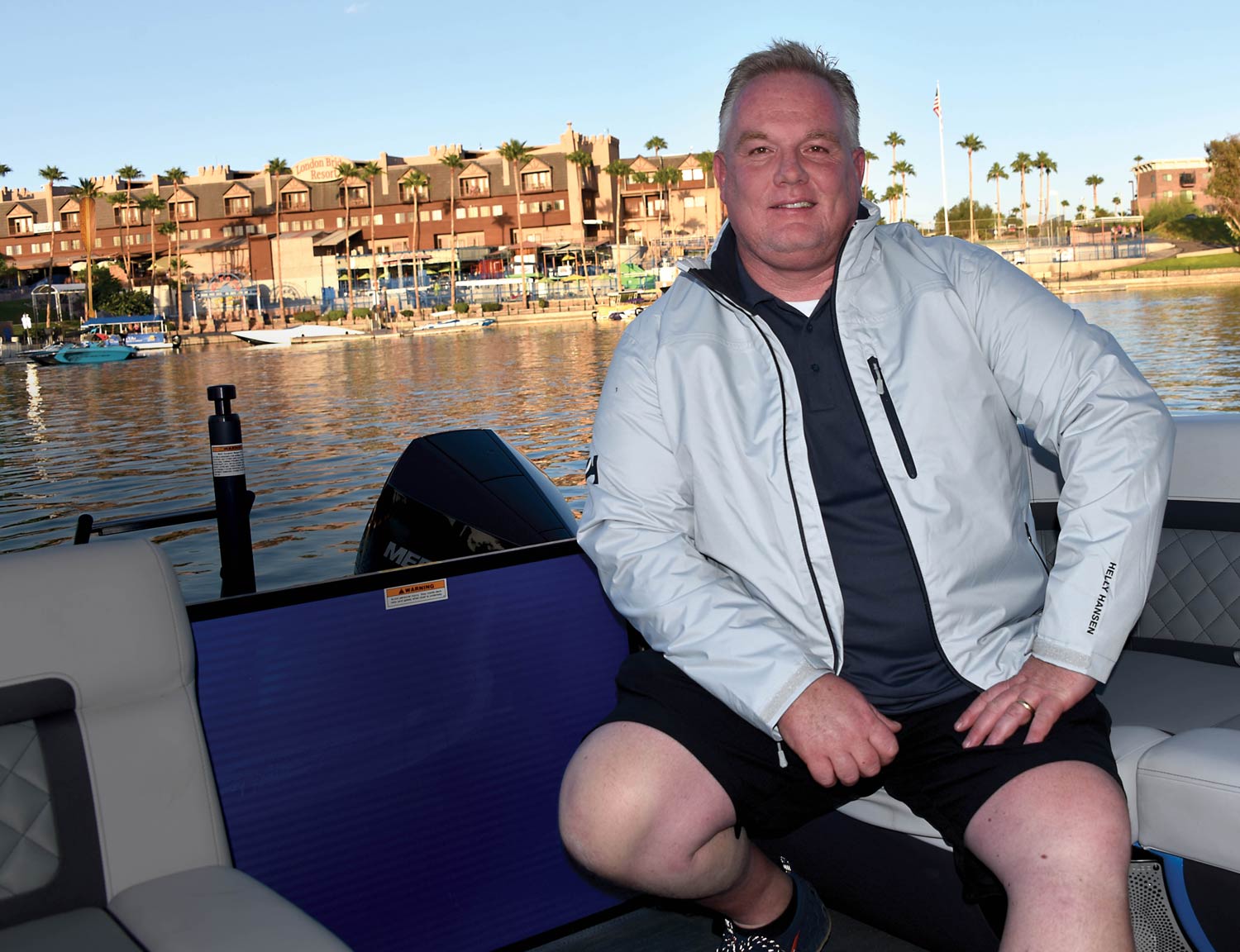 Brady L. Kay smiles and poses for a photograph outdoors on a clear sunny day as he sits back on a grey sofa couch inside a motorboat vehicle wearing a grey Helly Hansen branded crew midlayer sailing jacket