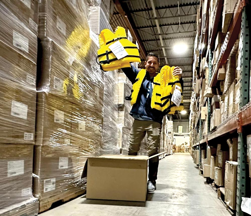 a man wearing a bright yellow life jacket and smiles at the camera as he removes more bright yellow life jackets from a cardboard box in a large factory