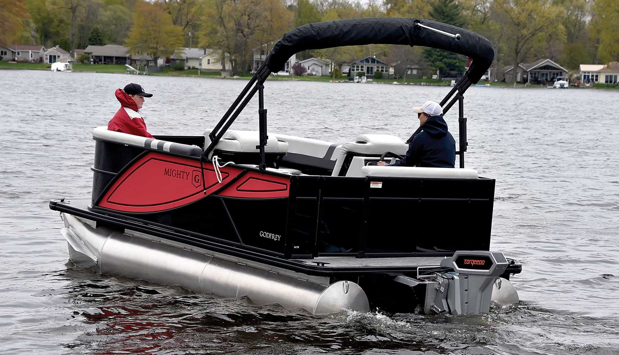 3/4ths rear view of a woman operating a Godfrey Mighty G series electric powered pontoon from the captains chair as a young boy relaxes on a bench seat