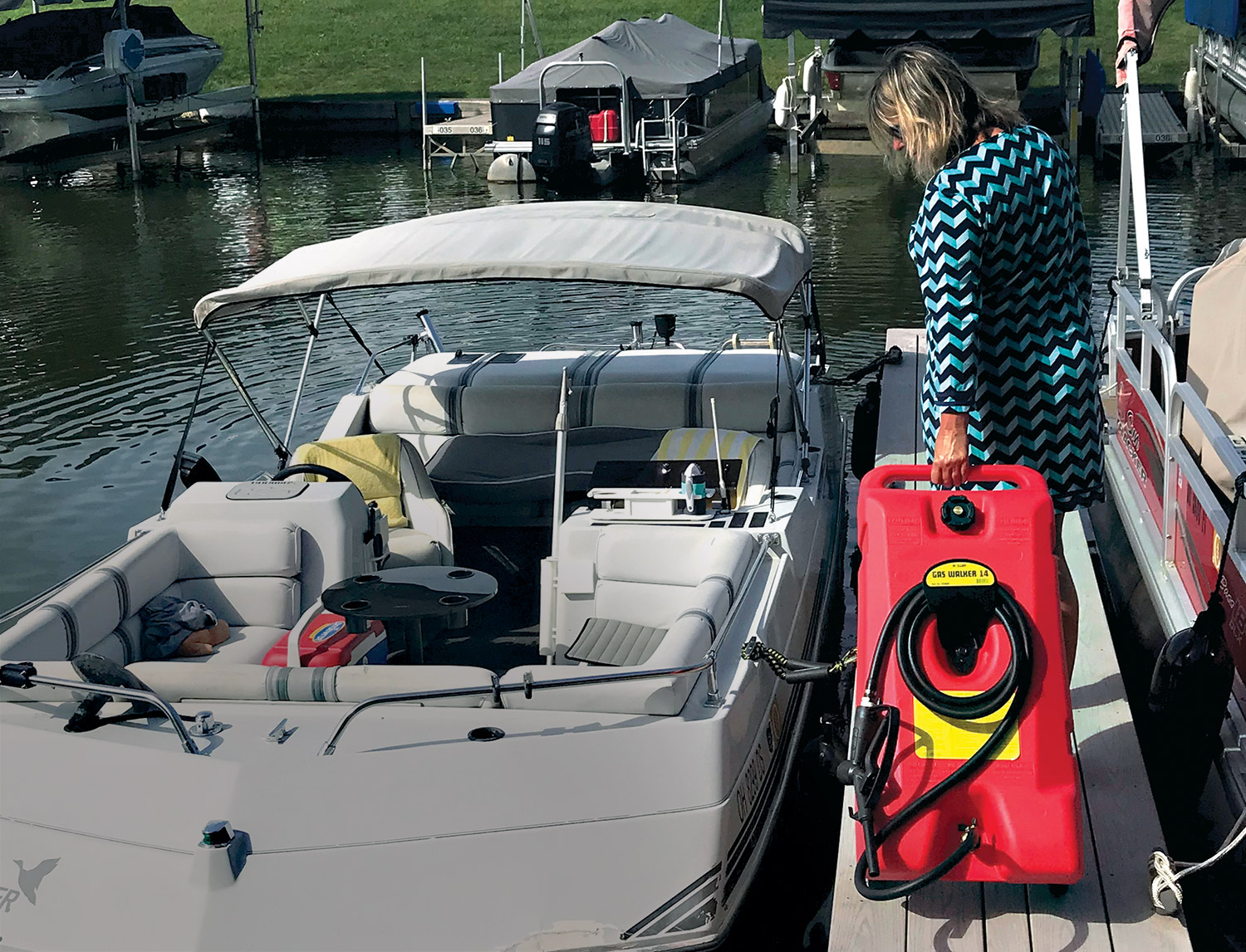 A woman on a dock landing area carries/drags the red/black/yellow colored Moeller Gas Walker 14 Portable Quick-Fill Fuel Station tank to the family deck boat beside her