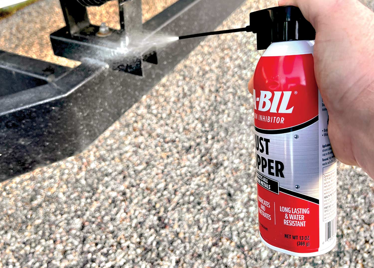 Close-up photograph perspective of person's hand gripping/spraying STA-BIL's Rust Stopper aerosol protectant onto a metal area of a boat trailer that contains some rust damage