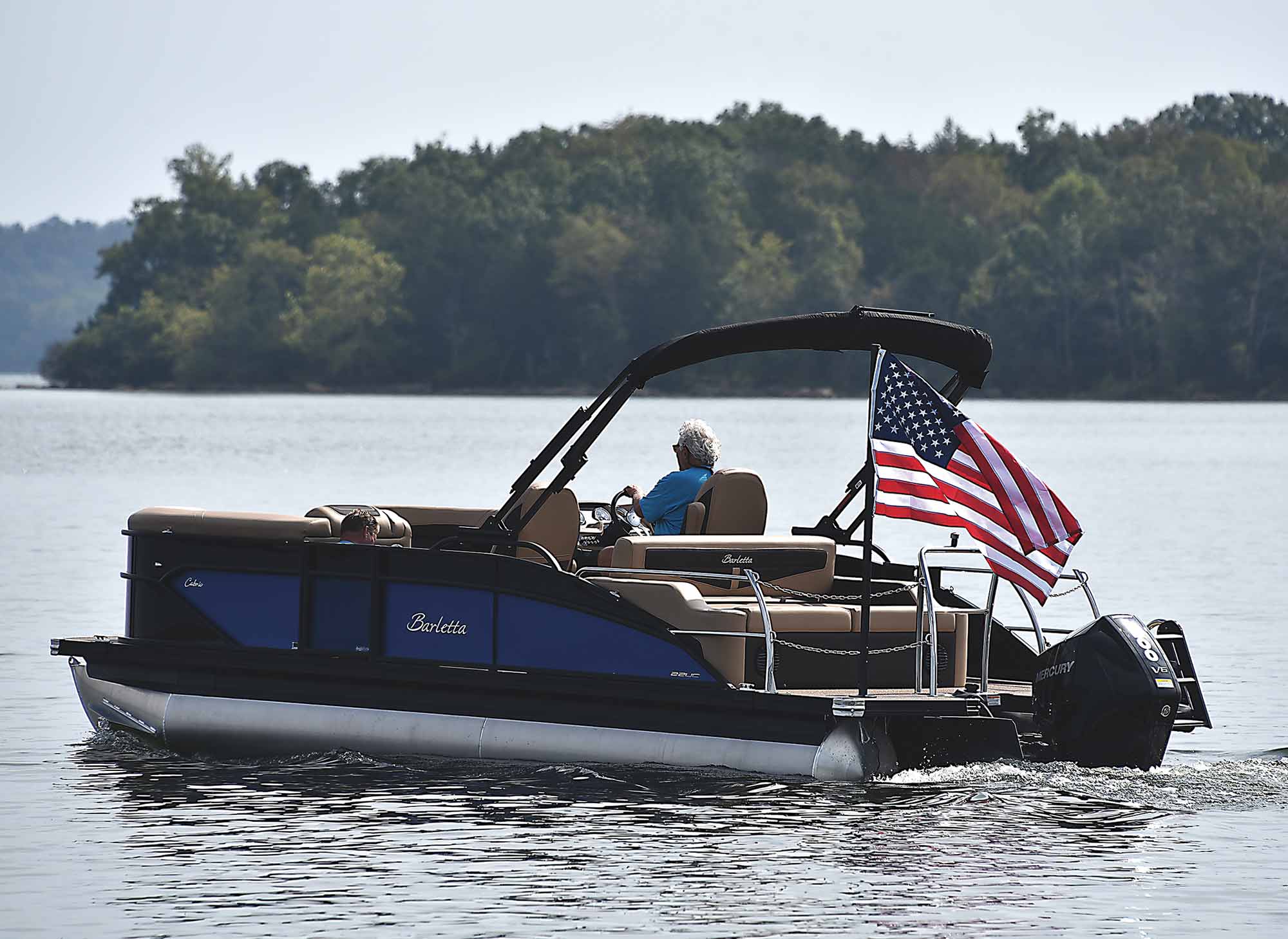 Landscape close-up photograph perspective of a man in a blue t-shirt and sunglasses driving a blue/black Barletta pontoon boat motor vehicle on the water with another passenger inside the boat plus a small American flagpole mounted stand (Lillipad Marine Flagpole/Grill Post) showing the United States of America flag waving in the air attached to the rear stern surface area of the boat