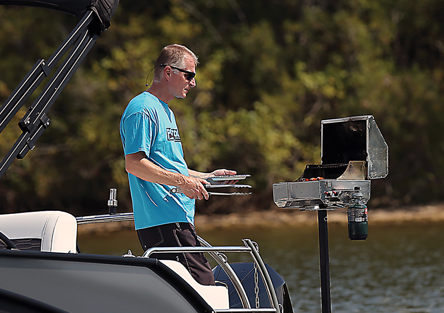 Close-up landscape photograph perspective of a man in a blue graphic t-shirt and sunglasses holding a stainless steel silver cooking tong plus a plastic plate in his hands while he is standing on the rear stern surface area of the pontoon boat motor vehicle glancing downward at the open small metal chrome colored mounted barbecue grill stand (Lillipad Marine Flagpole/Grill Post)