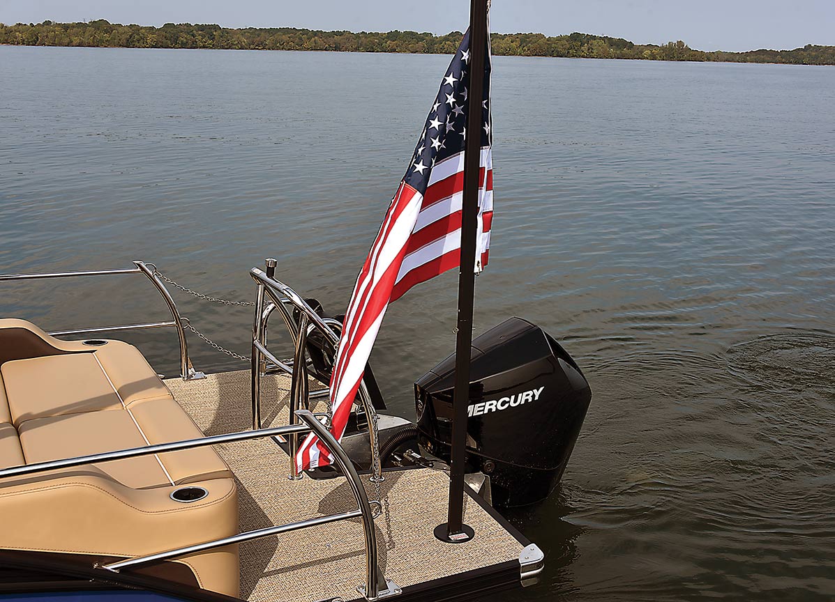 Close-up landscape photograph perspective of a small American flagpole mounted stand (Lillipad Marine Flagpole/Grill Post) showing the United States of America flag waving slightly in the air attached to the rear stern surface area of the pontoon boat motor vehicle