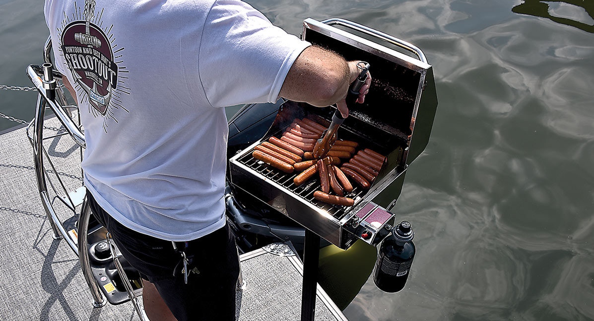 Close-up landscape photograph rear back perspective of a man in a white 2024 Pontoon and Deck Boat Shootout graphic t-shirt holding a stainless steel silver cooking tong as he cooks hot dog weenies on a open small metal chrome colored mounted barbecue grill stand (Lillipad Marine Flagpole/Grill Post) attached to the rear stern surface area of the pontoon boat motor vehicle