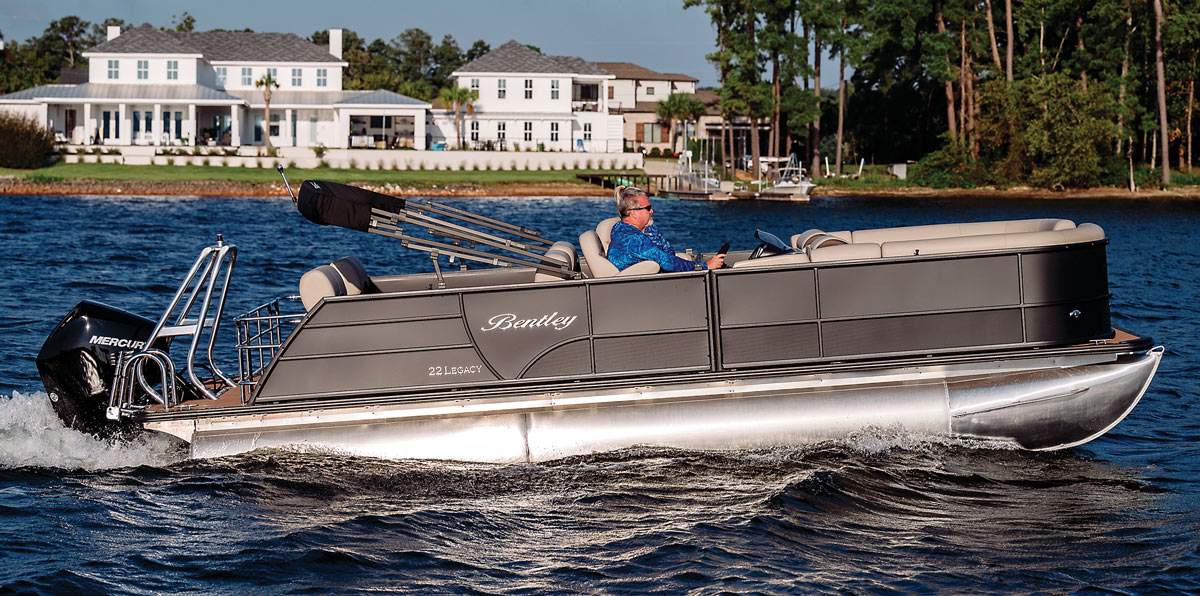 A man in a blue jacket driving a grey Bentley pontoon in front of two white houses and tree.