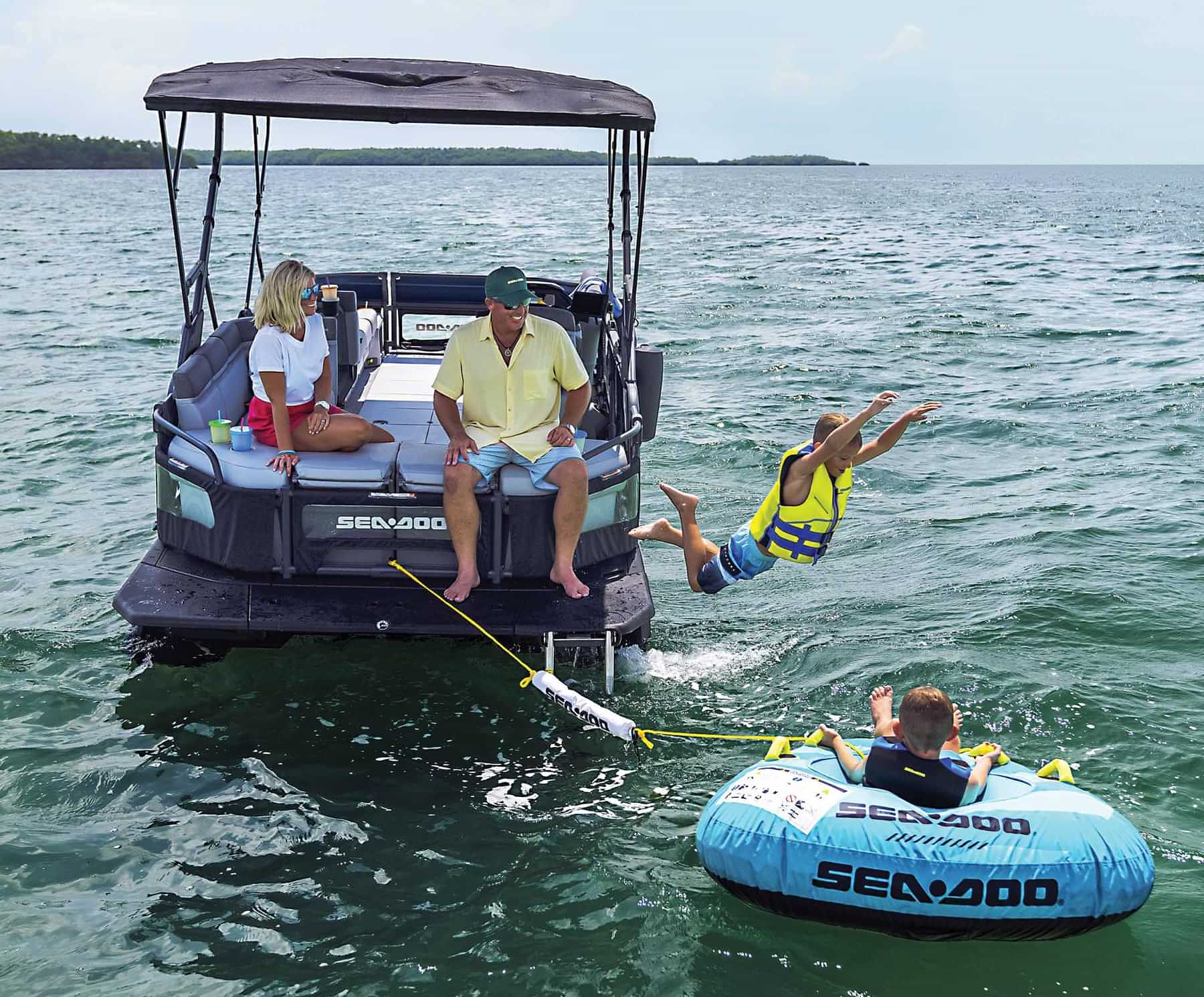a man and woman smile and watch from the rear seating of a Sea-Doo pontoon as a young boy wearing a life jacket jumps into the water next to another young boy, also wearing a life jacket and relaxing in a Sea-Doo inner tube tethered to the rear of the pontoon