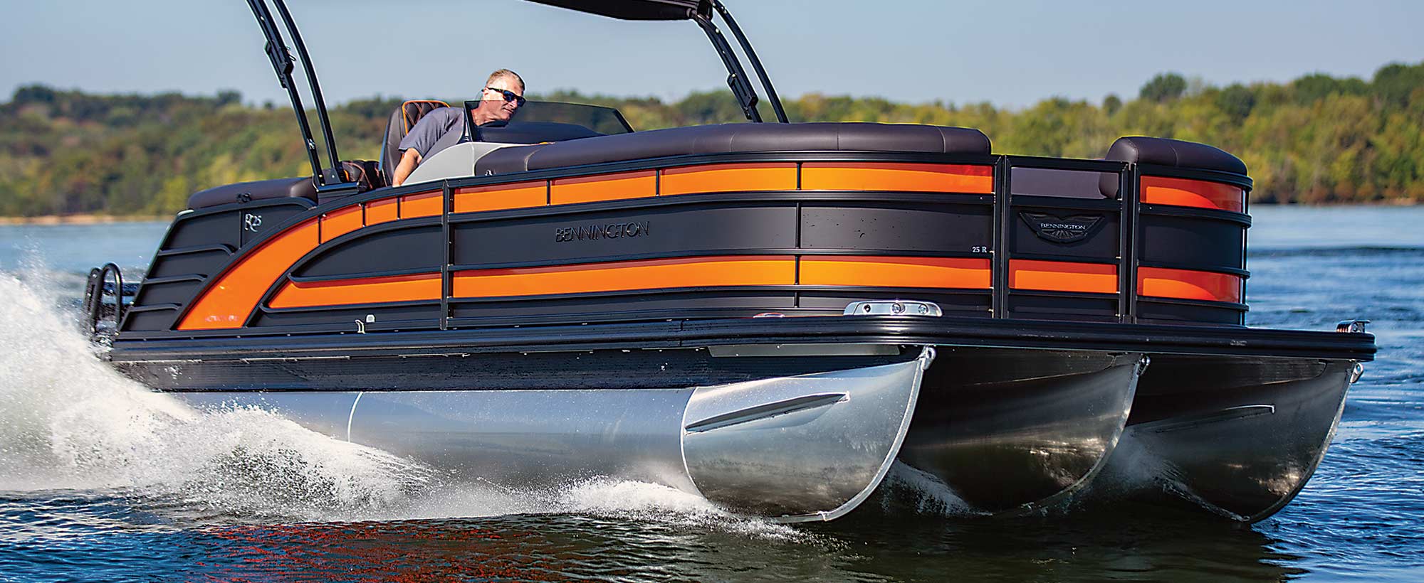 Man driving an orange and black Bennington sport pontoon with trees in the background