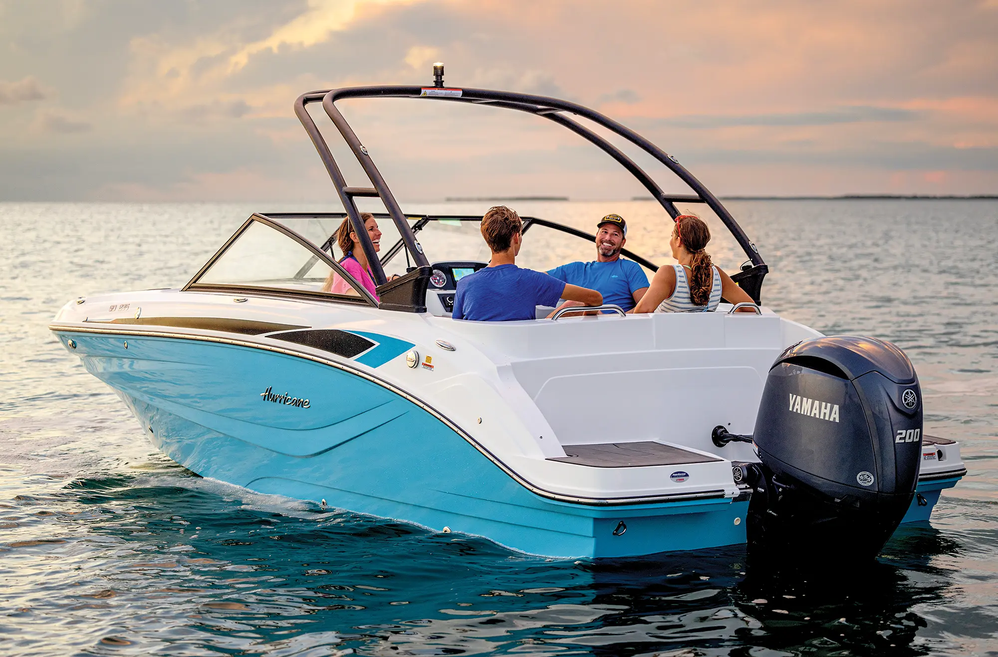 Aerial angle side photo view of the Hurricane SunDeck 235 pontoon motorboat vehicle as a woman, man, a younger boy, and younger girl are all seen smiling and laughing in their beach attire as they all talk amongst each other on a beautiful evening day