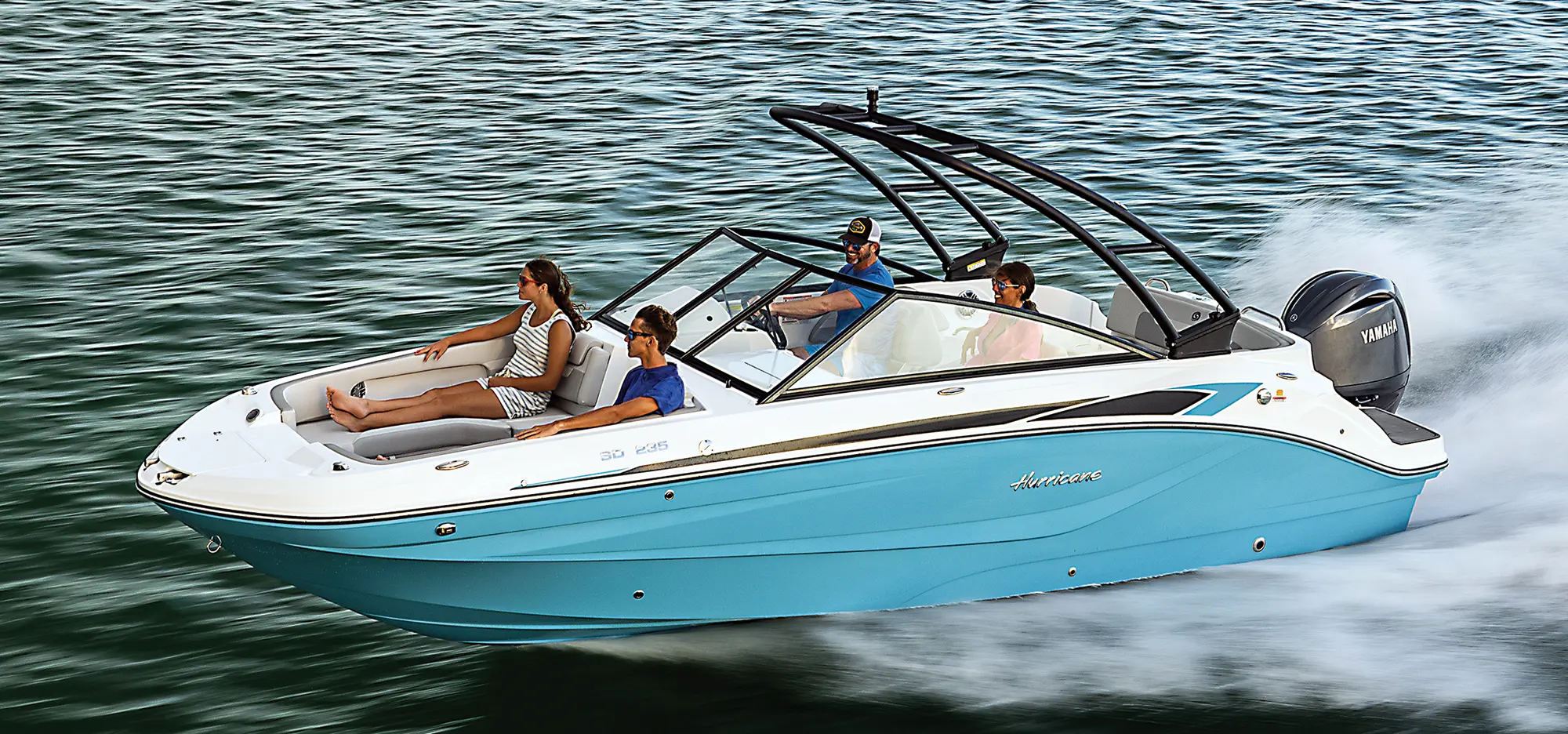 Aerial angle photo view of a man, woman, a younger girl, and younger boy sitting nearby each other inside the Hurricane SunDeck 235 pontoon motorboat vehicle as all of them are smiling in beach attire and sunglasses as the man in the hat is driving