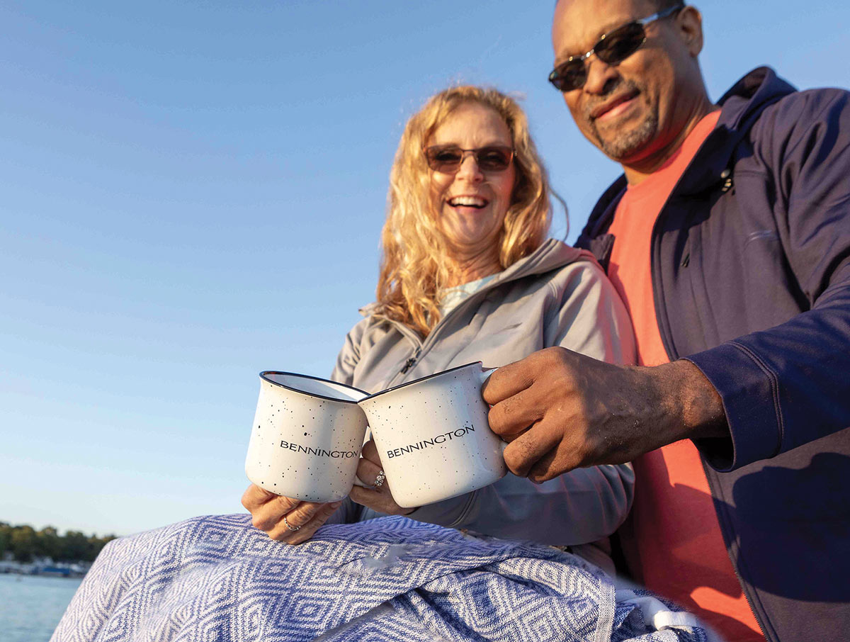 a couple relaxing near a body of water smile while holding their two Bennington mugs next to each other