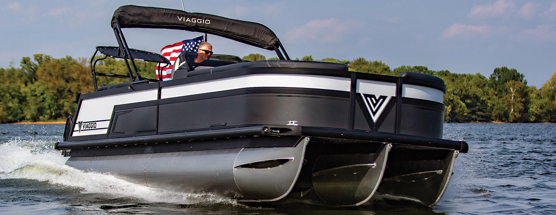 starboard side view of a man wearing sunglasses, operating a black Viaggio Diamante 26U pontoon on an enclosed body of water