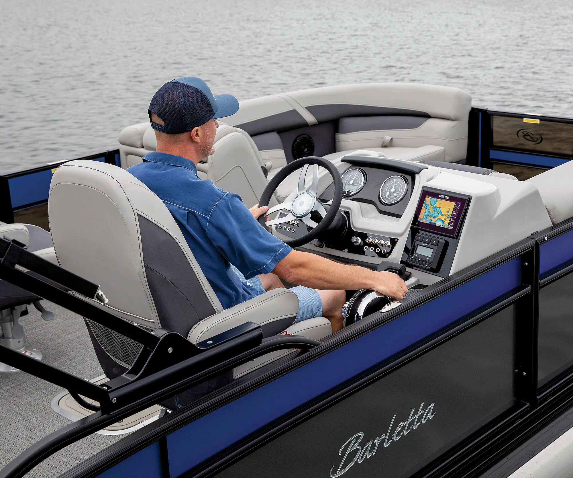 Landscape close-up photograph of a man in a dark blue button-up shirt and dark blue snapback hat in the driver's seat driving the Barletta Cabrio C22UC pontoon motorboat vehicle out in the water
