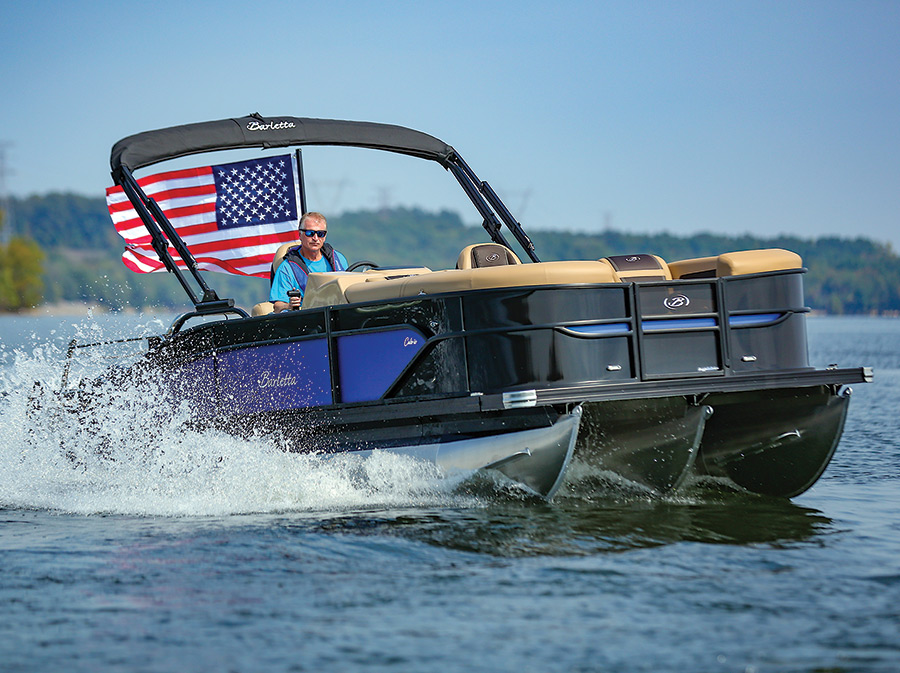 Landscape close-up photograph of a man in a bright light blue t-shirt and wearing dark sunglasses in the driver's seat driving the Barletta Cabrio C22UC pontoon motorboat vehicle out in the water as the motorboat vehicle has an American flag waving in motion at the back of the pontoon