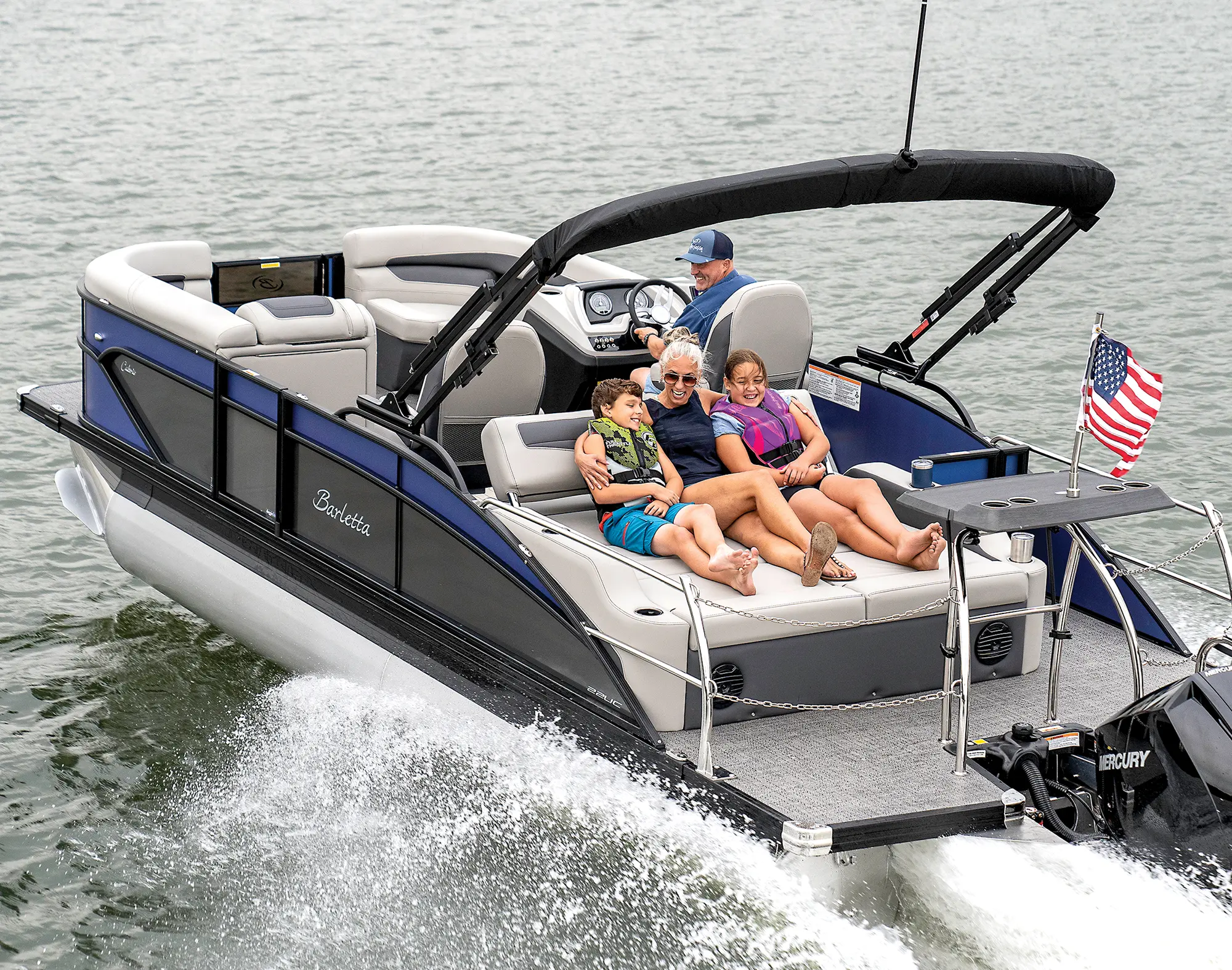 Landscape close-up photograph of a man in a dark blue button-up shirt and dark blue snapback hat in the driver's seat driving the Barletta Cabrio C22UC pontoon motorboat vehicle out in the water with a woman and children holding each other closely as they lean back and relax on a lounge couch chair
