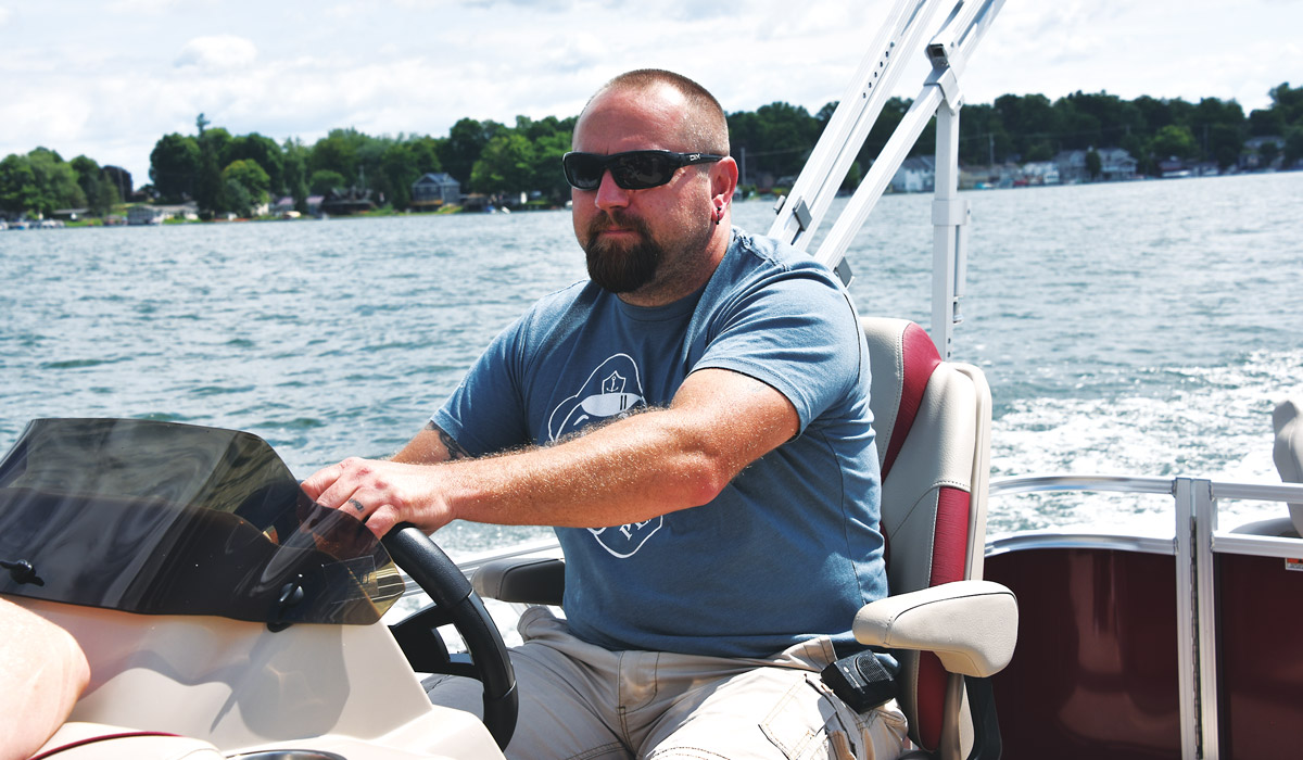 Man wearing sunglasses, a blue t-shirt, and tan shorts driving the Avalon Venture Cruise pontoon