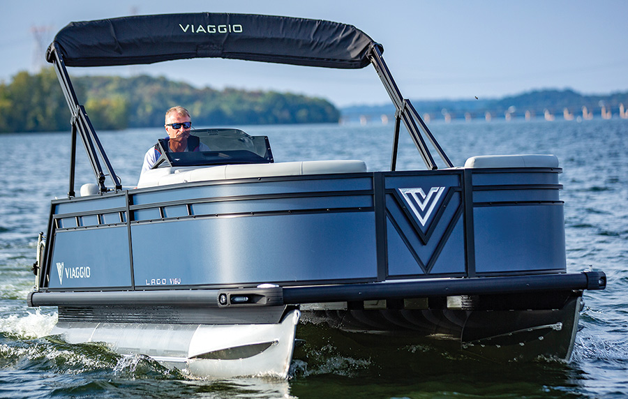 Landscape close-up side photograph view of a man in a t-shirt and sunglasses in the driver's seat driving the Viaggio Lago V16U pontoon motorboat vehicle out in the water