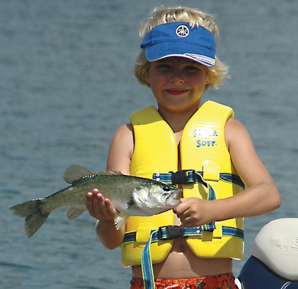 young boy with blonde hair wearing a blue visor and yellow life jacket holding a fish with both hands
