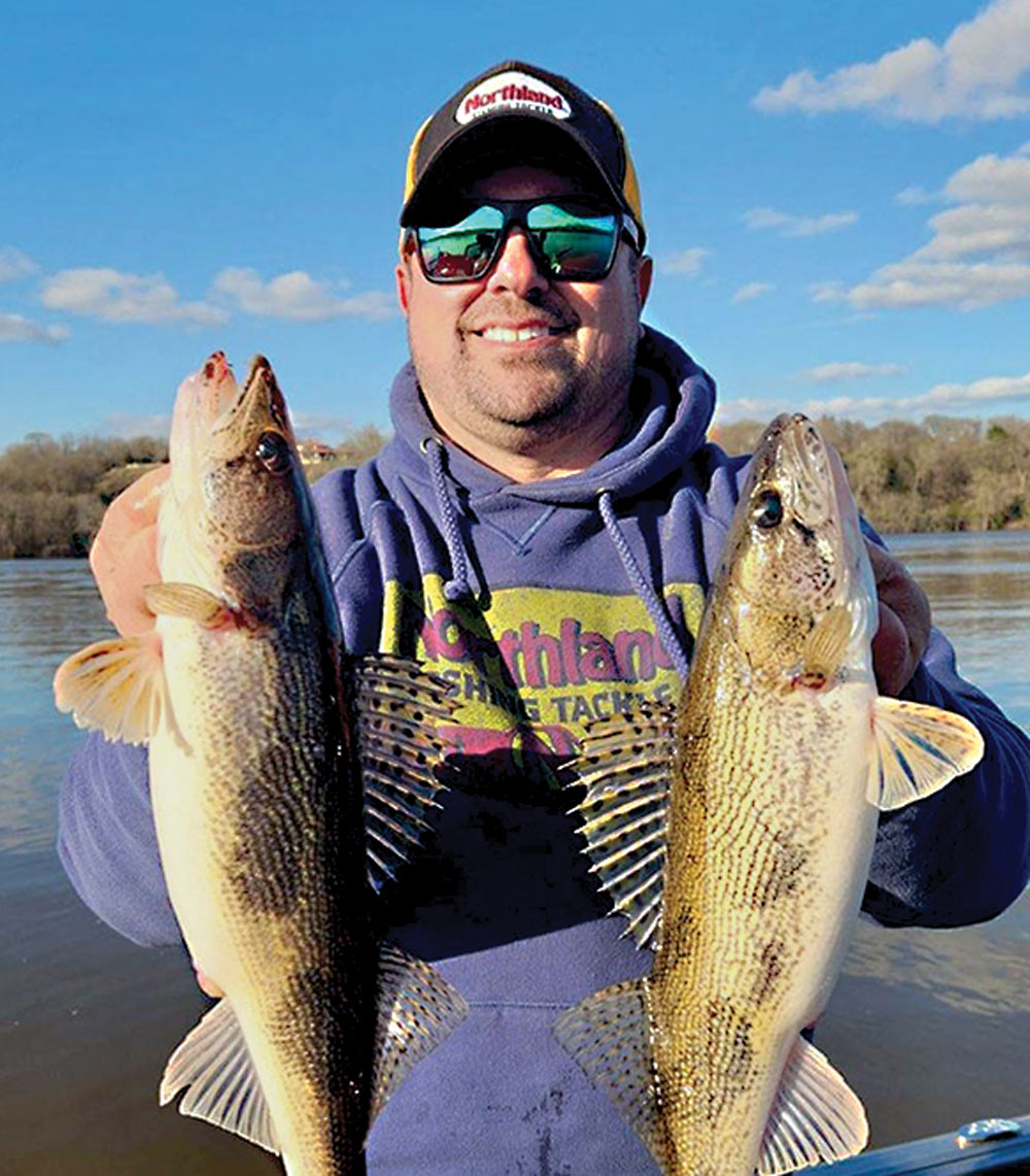 a man smiling while wearing sunglasses, a hat, and a blue sweater holding up two fish for a picture