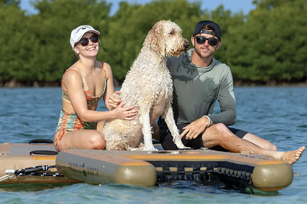 a woman, a man and a blonde curly haired medium to large sized dog sit on a larger floating pad beside an inflatable doggy pad connected to the larger pad by a rope