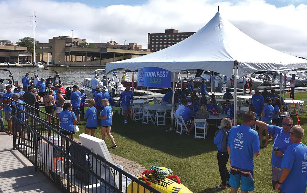 wide view of a 2022 ToonFest event tent pitched on a grassy area beside a city pier, many people dressed in summer clothes and blue event shirts stand and sit while mingling