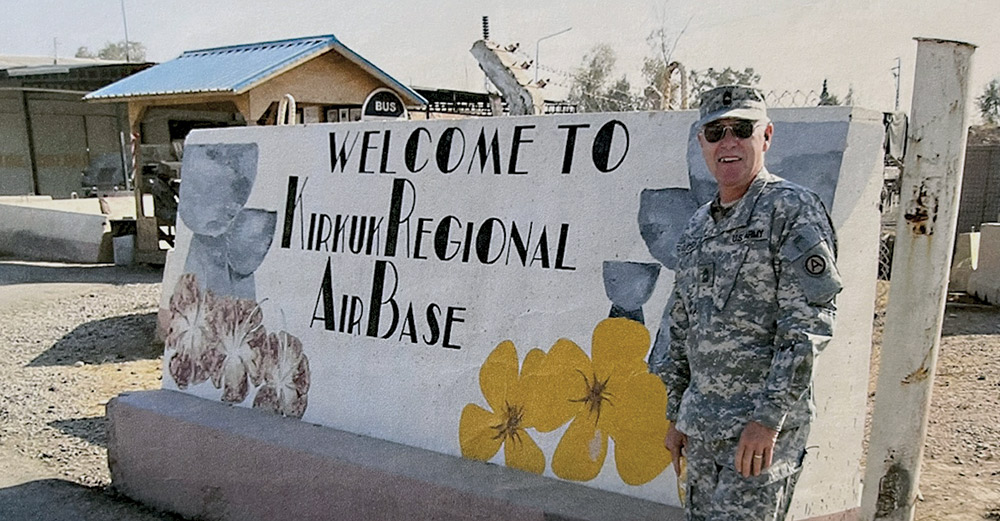 Landscape vintage close-up outdoor photograph view of Master Sergeant Joseph T. Houghton in his U.S. Army military outfit smiling as he poses in his sunglasses and military cap equipped while he stands next to the Welcome To Kirkuk Regional Air Base sign on a clear sunny day