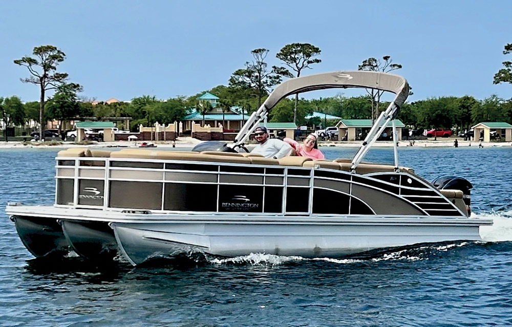 Landscape close-up angle outdoor photograph view of Aaron & Peyton Heintzelman in their beach swimwear attire smiling seated inside the dark brown/black Bennington Saltwater Series pontoon motorboat vehicle as Aaron is in the driver's seat and Peyton is seated on a passenger lounge chair while both of them are wearing sunglasses doing a funny silly pose as the pontoon is moving out on the water