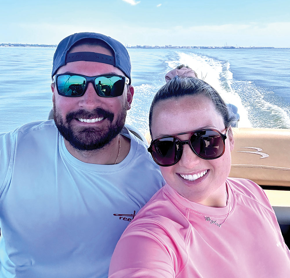 Portrait close-up outdoor selfie photograph view of Aaron & Peyton Heintzelman in their beach swimwear attire smiling inside the dark brown/black Bennington Saltwater Series pontoon motorboat vehicle as both of them are wearing sunglasses and Aaron is wearing a backwards snapback hat