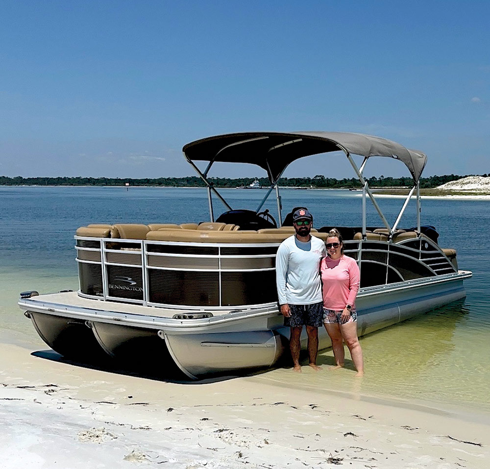 Portrait close-up angle outdoor photograph view of Aaron & Peyton Heintzelman in their beach swimwear attire smiling standing next to each other nearby the dark brown/black Bennington Saltwater Series pontoon motorboat vehicle as the pontoon is docked at the edge between the water and sand on a clear sunny day plus Aaron is wearing a hat