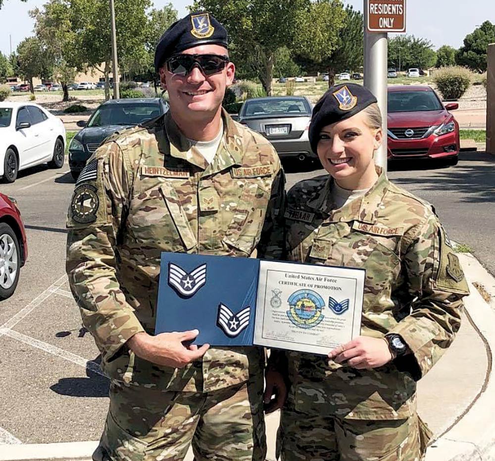 Portrait close-up outdoor photograph view of Aaron & Peyton Heintzelman in their United States Air Force military outfit smiling as they are holding together a small United States Air Force Certificate of Promotion rectangular shaped document book while they are standing next to each other in a parking lot area and Aaron is wearing sunglasses as well as both of them have on air force caps equipped on top of their head