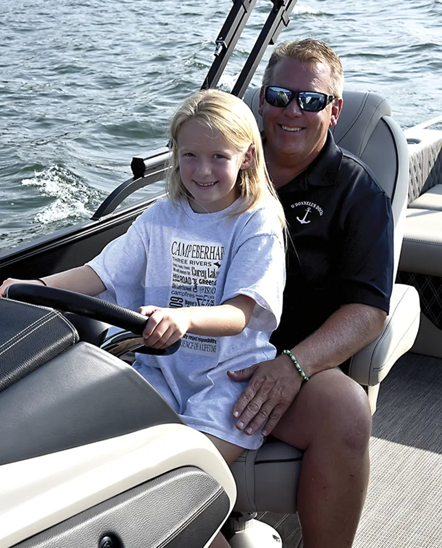 Shawn O'Donnell smiles while sitting at the helm of his black Barletta pontoon, his young daughter sits on his lap, smiling while grasping the wheel of the pontoon