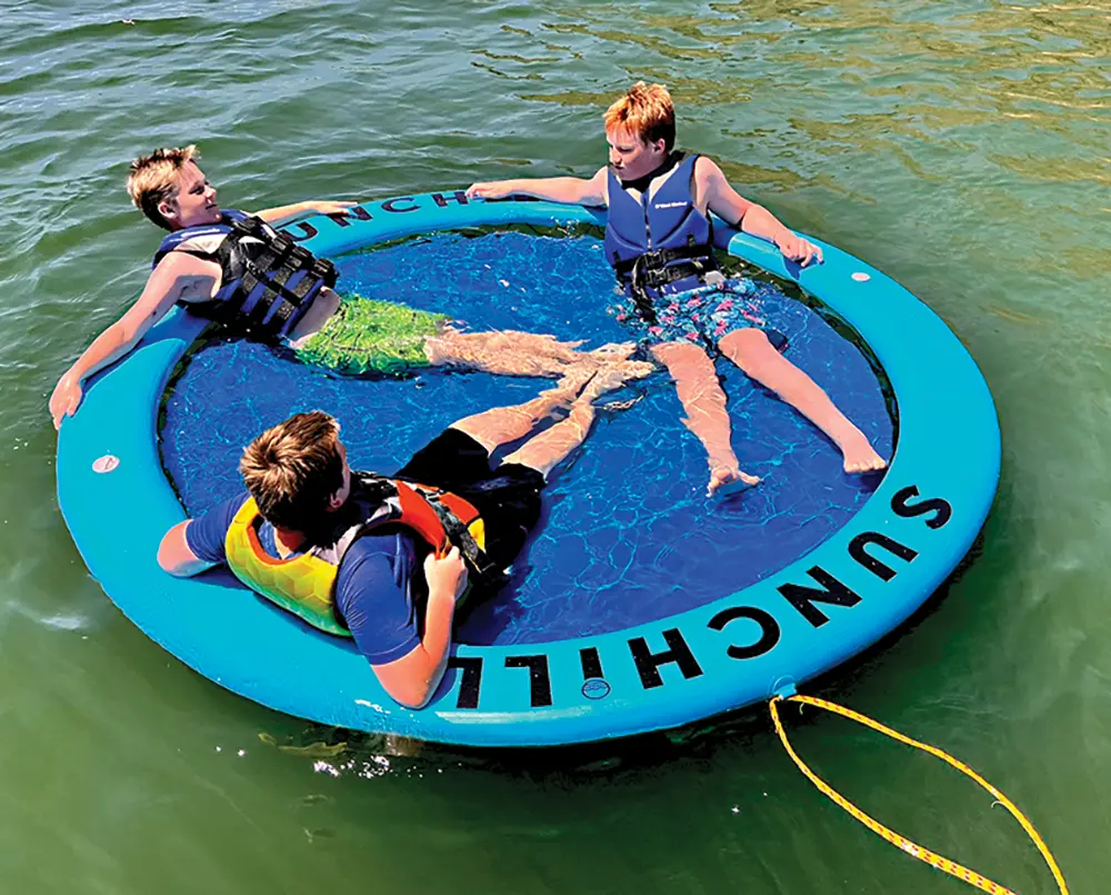 Portrait close-up photograph view of three male kids who are barefoot, have on tropical style pattern swimming shorts, and have on dark blue life swimming floatie vests/jackets as they are seen smiling/looking at each other with their arms stretched on top of the powder blue/dark blue Sunchill Water Hammock circular round shaped product model out in the water during the sunny day