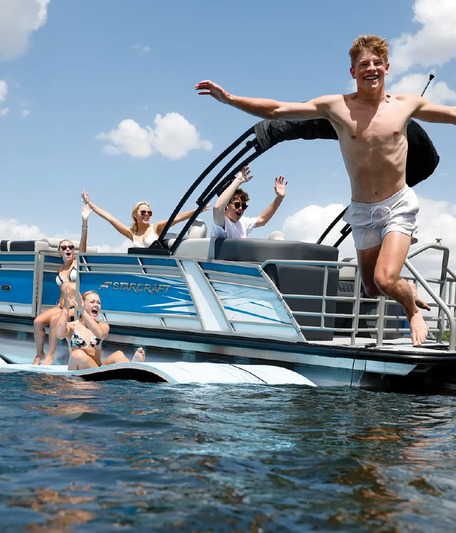 Portrait outdoor photograph of three young female individuals and two young male individuals all dressed up in beach attire as one of the male young individuals is seen smiling with his arms spread out and no shirt on/only shorts/barefoot in air in motion about to drop into the water as everyone else watches upon him as they all celebrate within proximity of a blue/white Starcraft Marine pontoon motorboat vehicle