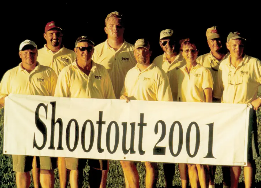 Vintage portrait photograph of nine Pontoon and Deck Boat Magazine staff members shown all posing for a group picture together while they are holding a white horizontal wide banner sign that says the phrase Shootout 2001 in black