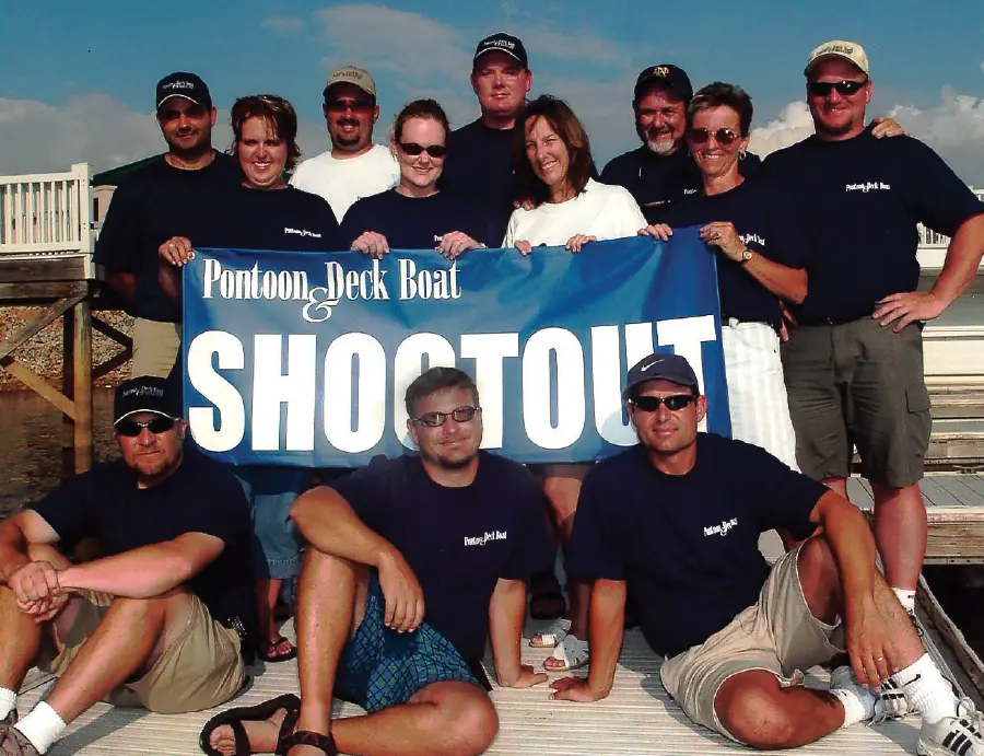 Vintage portrait photograph of twelve Pontoon and Deck Boat Magazine staff members shown all posing for a group picture together while they are holding a blue horizontal wide banner sign that says the phrase Pontoon & Deck Boat Shootout in white