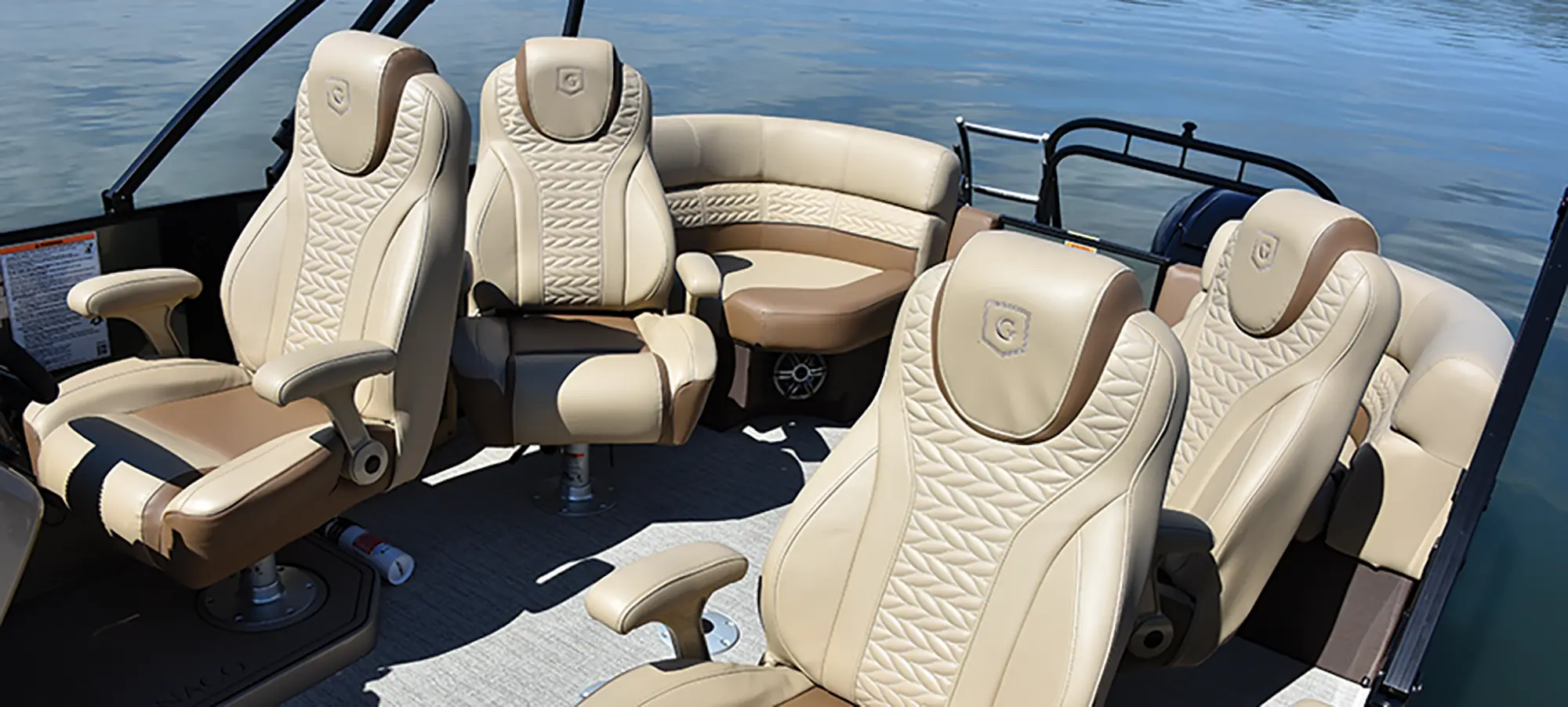 Angle landscape photograph of the Godfrey Monaco 235 QS pontoon motorboat vehicle featuring the tan/beige/dark brown driver's seat, three other leisure passenger seats, and lounge couch