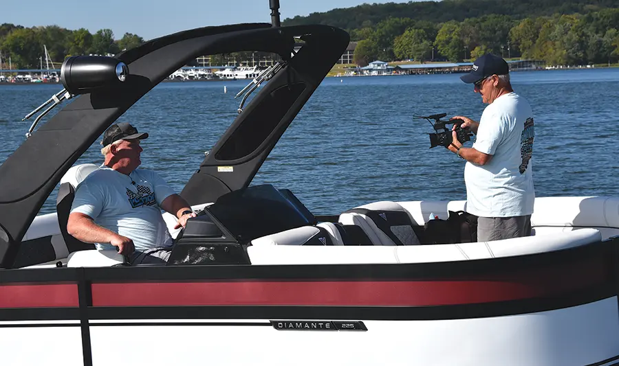 Landscape close-up photograph of Brady L. Kay in a light sky blue 2025 Pontoon and Deck Boat Shootout graphic t-shirt and a dark grey Pontoon typography hat seated down inside a red/black/white Viaggio Pontoons Diamante 225 pontoon motorboat vehicle model as he is glancing over at a video camera opposite another man standing in the same identical shirt and hat combination outfit who represents as a videographer recording footage of Brady L. Kay and is a staff member of Pontoon and Deck Boat as they are cruising in the water outdoors