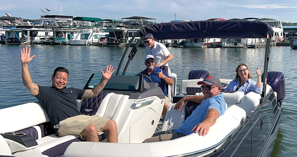 Five people smiling and waving on a pontoon boat at a marina.