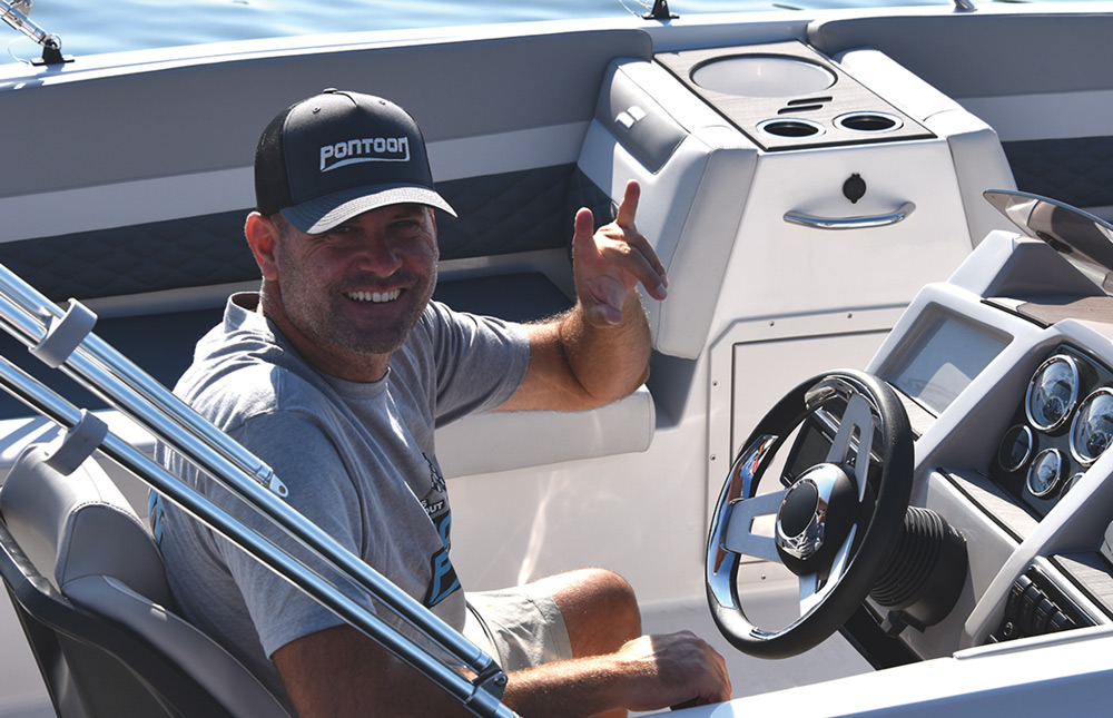Man sitting in a boat, smiling, with a black cap and gray t-shirt, steering wheel and dashboard visible.