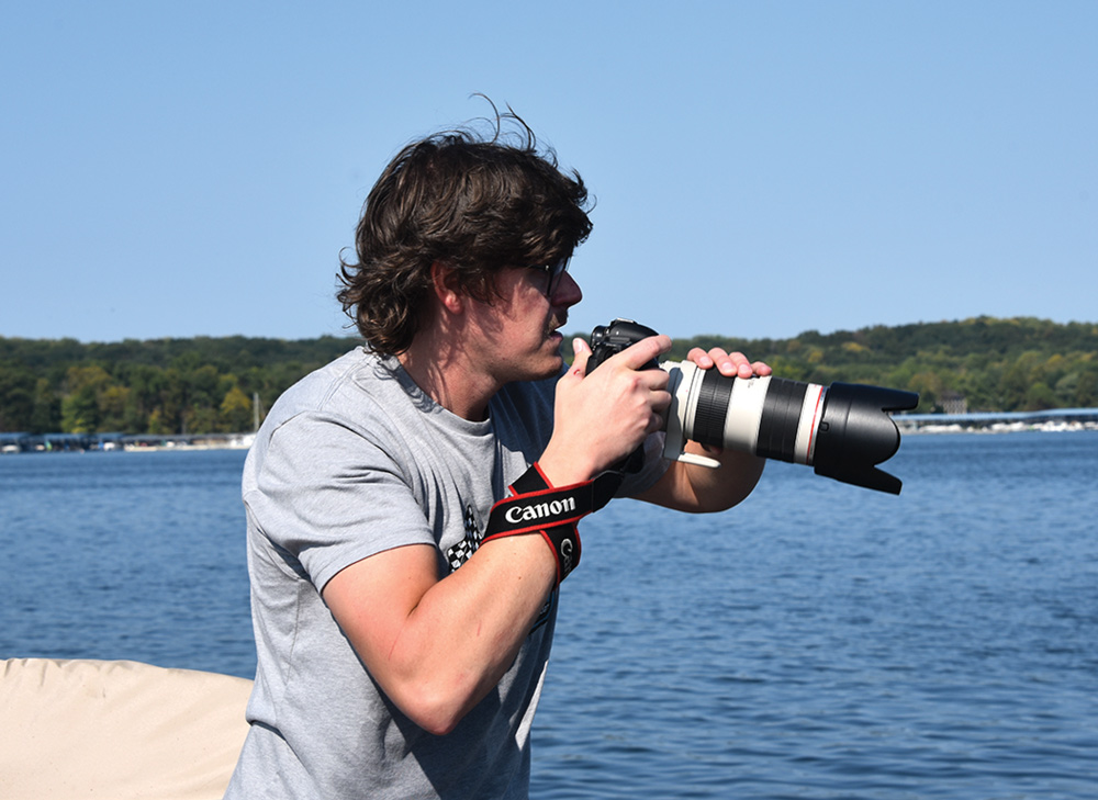 Person holding a camera with a long lens near a lake under a clear sky.