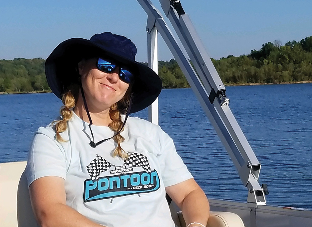 Woman wearing a hat and sunglasses sitting on a boat, with water and trees in the background.