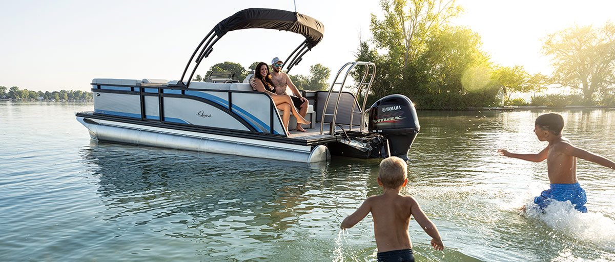 Pontoon boat with people onboard and children playing in the water nearby on a sunny day.