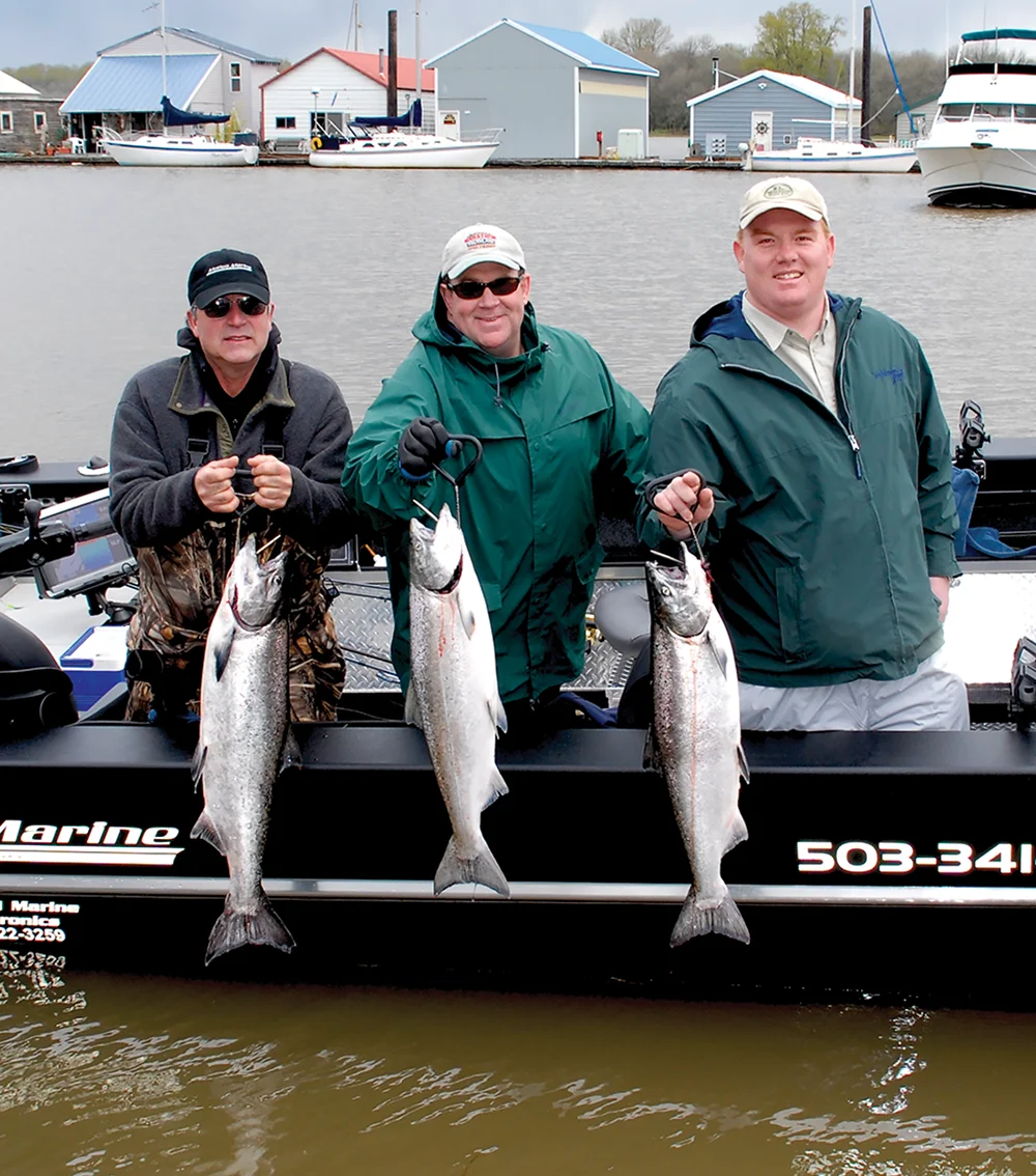 Three men holding large fish on a boat with other boats and buildings in the background.